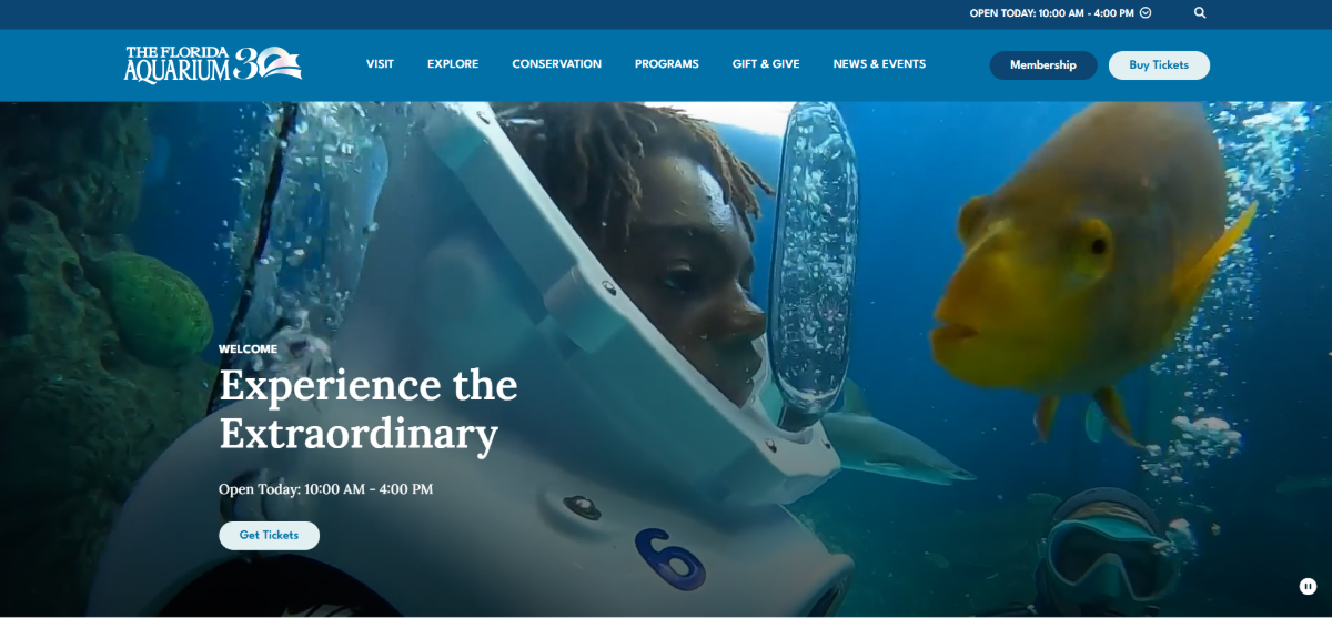 The homepage hero section for the Florida Aquarium website. We see a screenshot of the video section. There’s a kid in a deep-sea diving suit next to a large yellow fish. Over the video background, the text reads “Welcome: Experience the Extraordinary”. The aquarium’s hours of operation appear (10 a.m. to 4 p.m.) as well as a “Get Tickets” button.