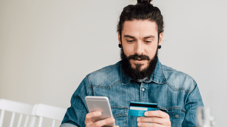 Man in a denim shirt looks at a smartphone while holding a credit card, suggesting an online payment or shopping transaction.